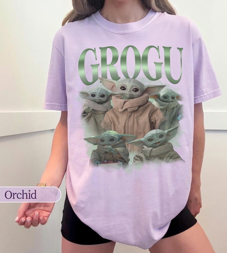 Op de afbeelding: Een lichtpaars t-shirt met het woord "GROGU" in groene metallic letters. Het shirt toont een afbeelding van Grogu, ook bekend als Baby Yoda, uit de Star Wars-serie. Het t-shirt is lichtpaars van kleur.