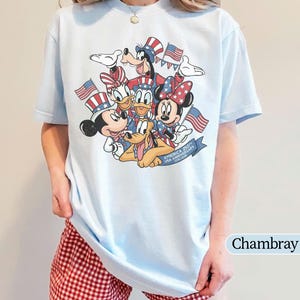 May include: Light blue t-shirt featuring a graphic of Disney characters Mickey Mouse, Minnie Mouse, Donald Duck, Daisy Duck, Goofy, and Pluto, with American flags and text "America 250th USA Anniversary 1776-2026".