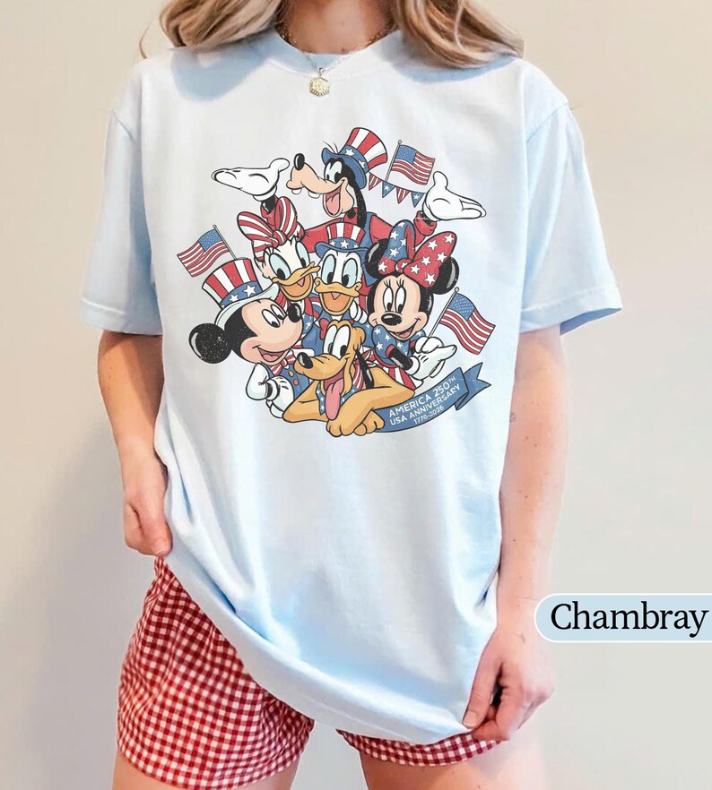 May include: Light blue t-shirt featuring a graphic of Disney characters Mickey Mouse, Minnie Mouse, Donald Duck, Daisy Duck, Goofy, and Pluto, with American flags and text "America 250th USA Anniversary 1776-2026".
