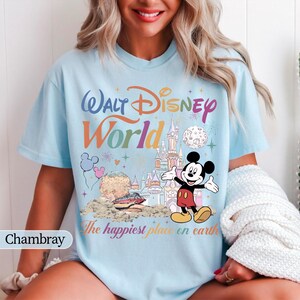 May include: Light blue t-shirt with a colorful "Walt Disney World" graphic featuring Mickey Mouse, a castle, and the phrase "The happiest place on earth." The design includes balloons and fireworks.