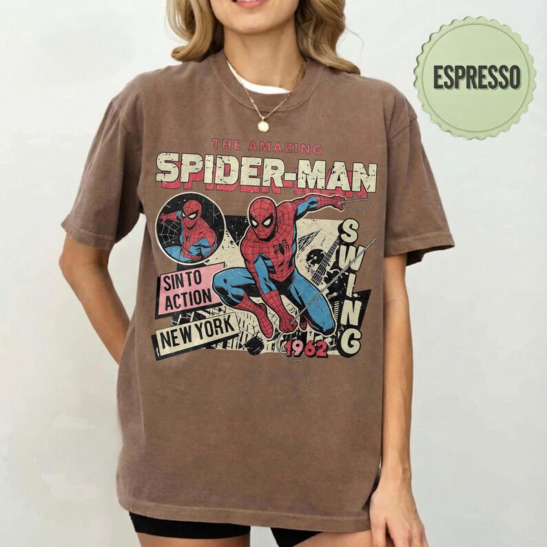 Op de afbeelding: Bruin t-shirt met een vintage stripboekafbeelding van Spider-Man. Het ontwerp bevat de tekst "THE AMAZING SPIDER-MAN", "SIN TO ACTION" en "NEW YORK 1962". Het shirt heeft een ronde hals en korte mouwen.