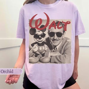 May include: Orchid-colored t-shirt featuring a sepia-toned graphic of Mickey Mouse wearing sunglasses next to Walt Disney. The word "Walt" is printed in red script above the image. The shirt has short sleeves and a relaxed fit.