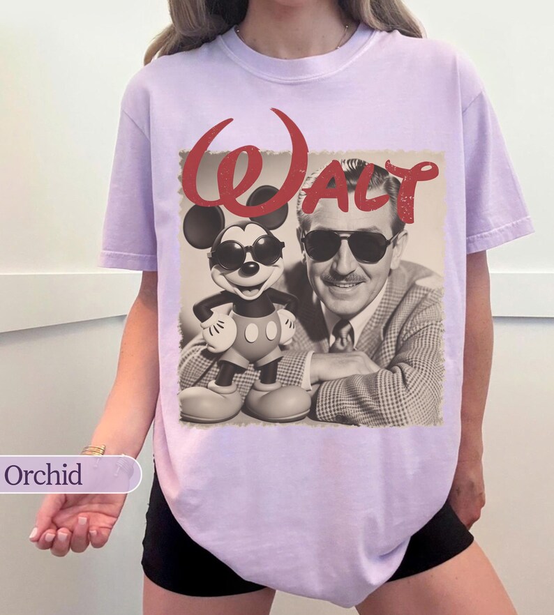 May include: Orchid-colored t-shirt featuring a sepia-toned graphic of Mickey Mouse wearing sunglasses next to Walt Disney. The word "Walt" is printed in red script above the image. The shirt has short sleeves and a relaxed fit.