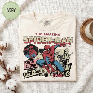 Op de afbeelding: Ivoor T-shirt met een vintage Spider-Man graphic. Het ontwerp bevat de tekst "THE AMAZING SPIDER-MAN", "SIN TO ACTION" en "NEW YORK", samen met een illustratie van Spider-Man. Het shirt is opgevouwen en gepresenteerd op een witte ondergrond.
