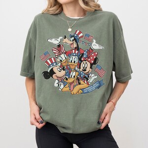 May include: Olive green t-shirt featuring a graphic of Mickey Mouse, Minnie Mouse, Donald Duck, Daisy Duck, Goofy, and Pluto with American flags and patriotic attire. The text reads "America 250th USA Anniversary".