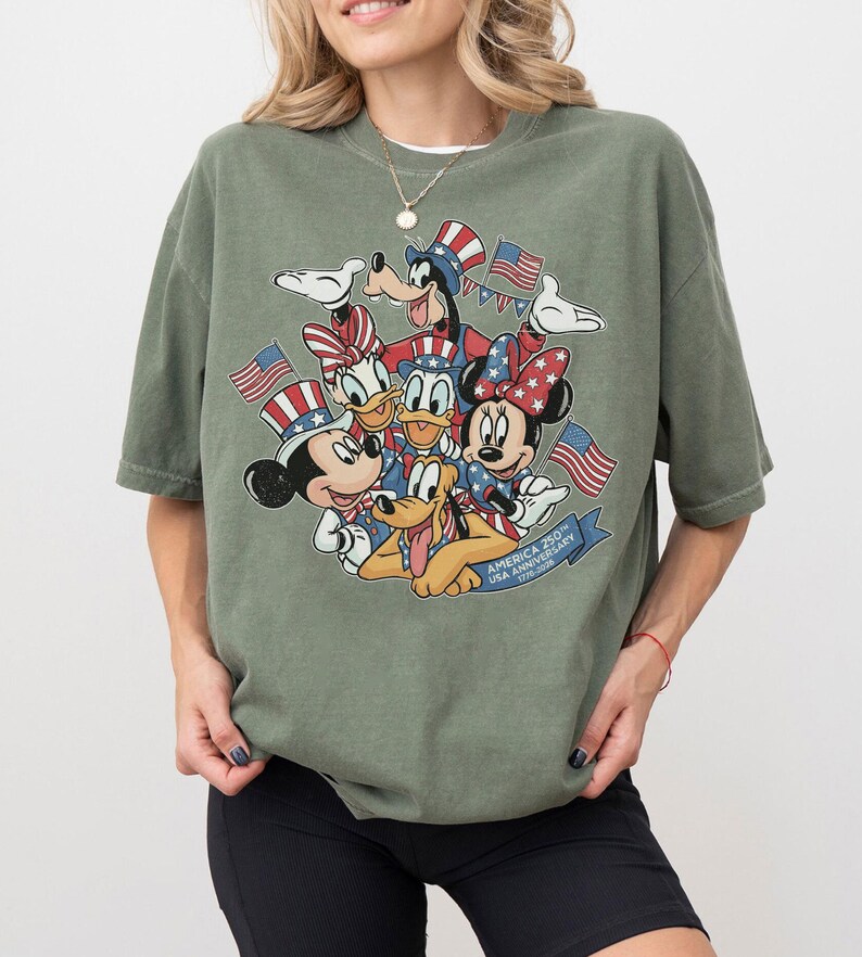 May include: Olive green t-shirt featuring a graphic of Mickey Mouse, Minnie Mouse, Donald Duck, Daisy Duck, Goofy, and Pluto with American flags and patriotic attire. The text reads "America 250th USA Anniversary".