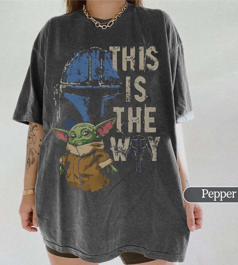 Może przedstawiać: Ciemnoszara koszulka z grafiką niebieskiego hełmu, zielonego kosmity i napisem "THIS IS THE WAY". Projekt ma wygląd vintage. Koszulka ma kr&oacute;tkie rękawy i luźny kr&oacute;j.