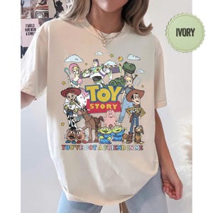 May include: Ivory-colored t-shirt featuring a colorful graphic of characters from the animated film "Toy Story." The design includes the text "TOY STORY" and "YOU'VE GOT A FRIEND IN ME." The shirt has a crew neck and short sleeves.