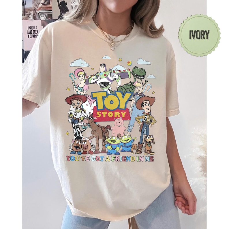 May include: Ivory-colored t-shirt featuring a colorful graphic of characters from the animated film "Toy Story." The design includes the text "TOY STORY" and "YOU'VE GOT A FRIEND IN ME." The shirt has a crew neck and short sleeves.