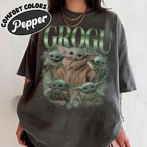 Op de afbeelding: Donkergrijs T-shirt met een afbeelding van Grogu uit The Mandalorian. Het woord "GROGU" is in het groen boven de afbeelding van Grogu gedrukt. Het shirt heeft ook de woorden "COMFORT COLORS" en "Pepper" erop staan.