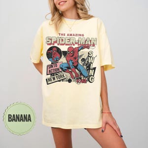 Op de afbeelding: Lichtgeel T-shirt met een Spider-Man afbeelding. Het ontwerp bevat de tekst "THE AMAZING SPIDER-MAN", "SINTO ACTION NEW YORK 1962". Het shirt heeft een losse pasvorm en is gemaakt van zacht materiaal.
