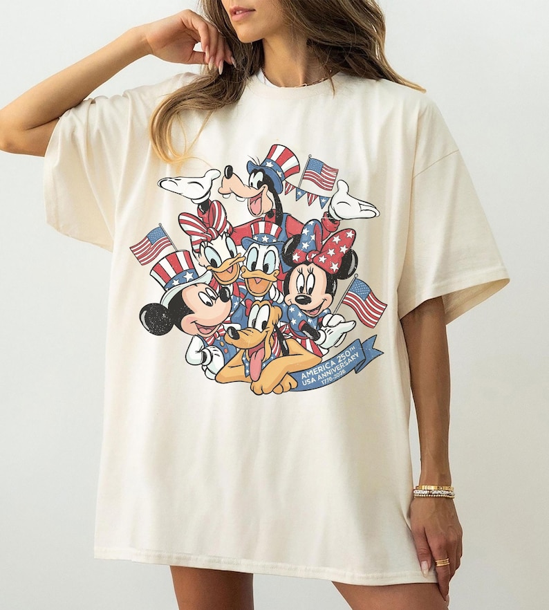 May include: Cream-colored t-shirt featuring a graphic of Mickey Mouse, Minnie Mouse, Donald Duck, Daisy Duck, Goofy, and Pluto in patriotic attire, holding American flags. The design includes the text "AMERICA 250 USA ANNIVERSARY 1776-2026".