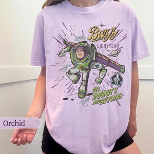 Puede incluir: Camiseta color orqu&iacute;dea con un gr&aacute;fico de Buzz Lightyear en una pose din&aacute;mica. El dise&ntilde;o incluye el texto "Buzz Lightyear" y "To Infinity and Beyond" con elementos espaciales. La camiseta es de uso casual y c&oacute;modo.