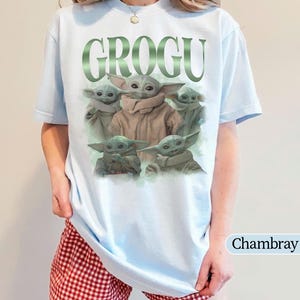 Op de afbeelding: Lichtblauw T-shirt met het woord "GROGU" in groen, met meerdere afbeeldingen van Grogu, ook bekend als Baby Yoda, uit de Star Wars-serie. Het shirt is lichtblauw van kleur en heeft een ronde hals.