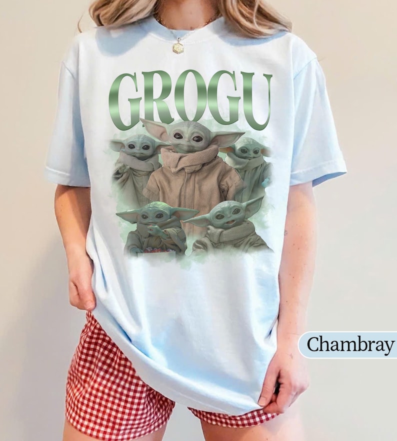 Op de afbeelding: Lichtblauw T-shirt met het woord "GROGU" in groen, met meerdere afbeeldingen van Grogu, ook bekend als Baby Yoda, uit de Star Wars-serie. Het shirt is lichtblauw van kleur en heeft een ronde hals.