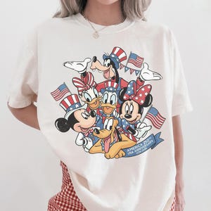May include: Off-white t-shirt featuring a graphic of Disney characters Mickey Mouse, Minnie Mouse, Donald Duck, Goofy, and Pluto, all in patriotic attire, holding American flags. The shirt commemorates the 250th USA Anniversary.