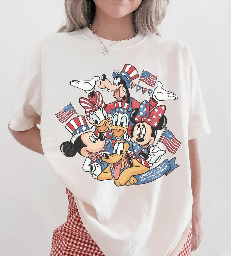 May include: Off-white t-shirt featuring a graphic of Disney characters Mickey Mouse, Minnie Mouse, Donald Duck, Goofy, and Pluto, all in patriotic attire, holding American flags. The shirt commemorates the 250th USA Anniversary.