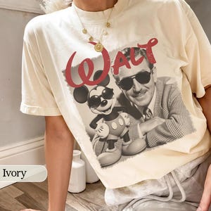 May include: Ivory-colored t-shirt featuring a graphic of Mickey Mouse and Walt Disney in sunglasses. The word "WALT" is printed in red above the image. The shirt is a relaxed fit.