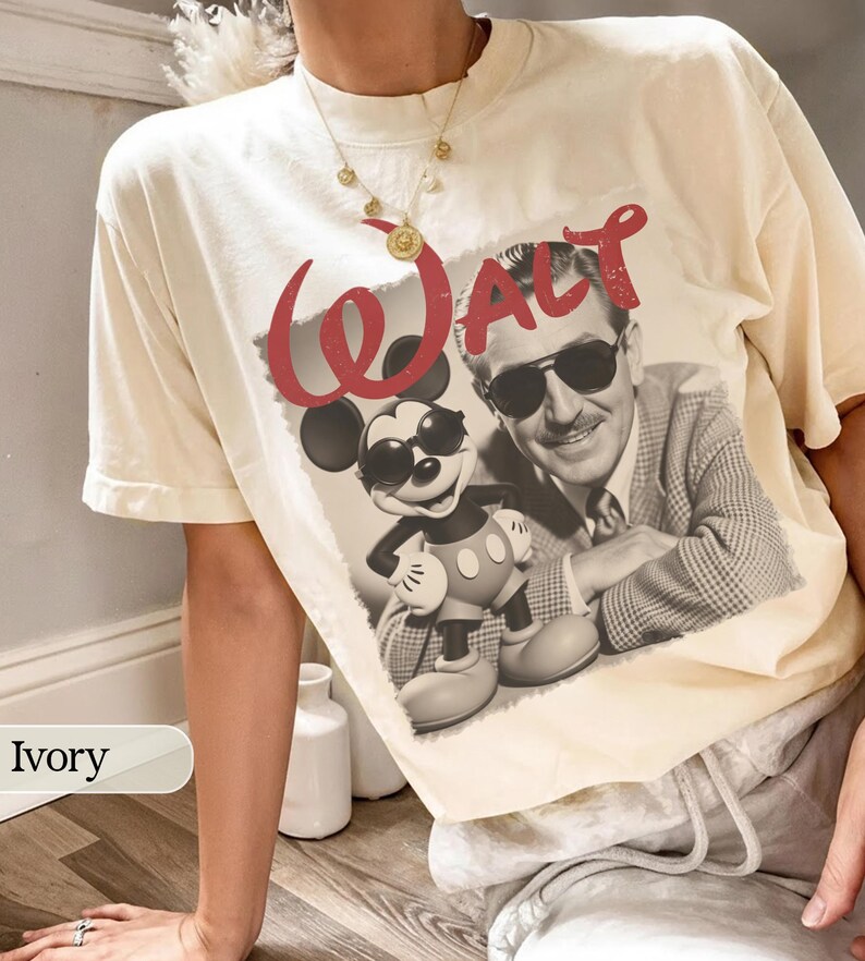 May include: Ivory-colored t-shirt featuring a graphic of Mickey Mouse and Walt Disney in sunglasses. The word "WALT" is printed in red above the image. The shirt is a relaxed fit.