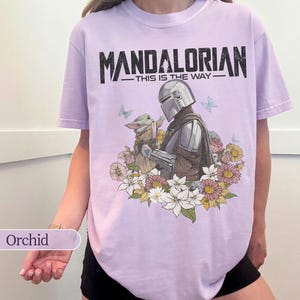 May include: Orchid-colored t-shirt featuring a graphic of The Mandalorian and Grogu surrounded by flowers and butterflies. The text "MANDALORIAN - THIS IS THE WAY" is printed across the top in black.