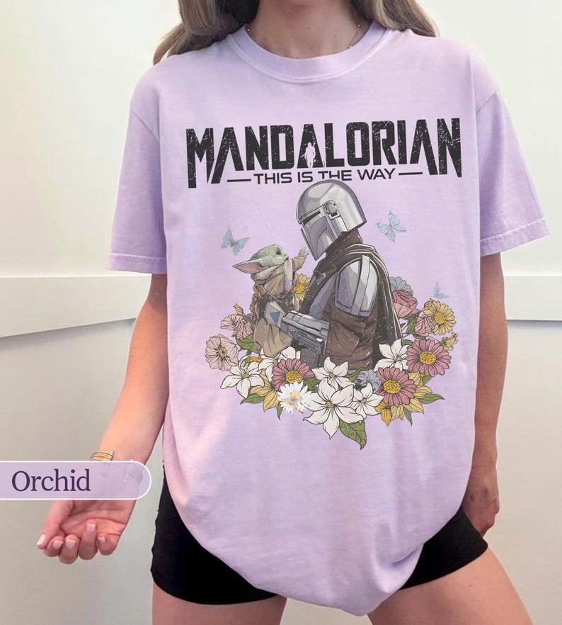 May include: Orchid-colored t-shirt featuring a graphic of The Mandalorian and Grogu surrounded by flowers and butterflies. The text "MANDALORIAN - THIS IS THE WAY" is printed across the top in black.