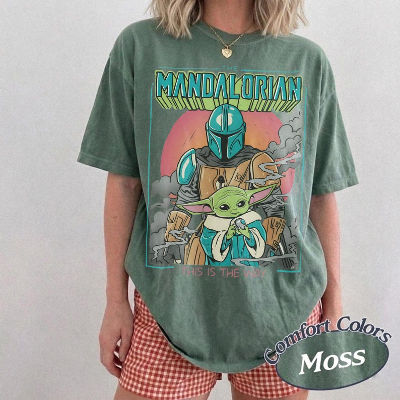 May include: A moss green short-sleeve t-shirt featuring a graphic print of The Mandalorian and Grogu. The text reads THE MANDALORIAN at the top and THIS IS THE WAY at the bottom. The design includes a sunset background with clouds and a teal helmet.