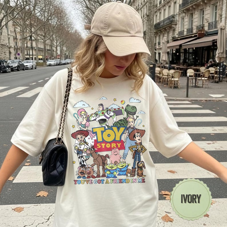 May include: Ivory-colored t-shirt featuring a colorful graphic of characters from the animated film "Toy Story." The design includes Woody, Buzz Lightyear, and other characters, with the text "TOY STORY" and "YOU'VE GOT A FRIEND IN ME."