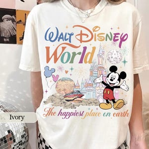 May include: Ivory-colored t-shirt featuring a colorful "Walt Disney World" graphic with Mickey Mouse, a castle, and the phrase "The happiest place on earth." The design includes balloons, fireworks, and a planet.