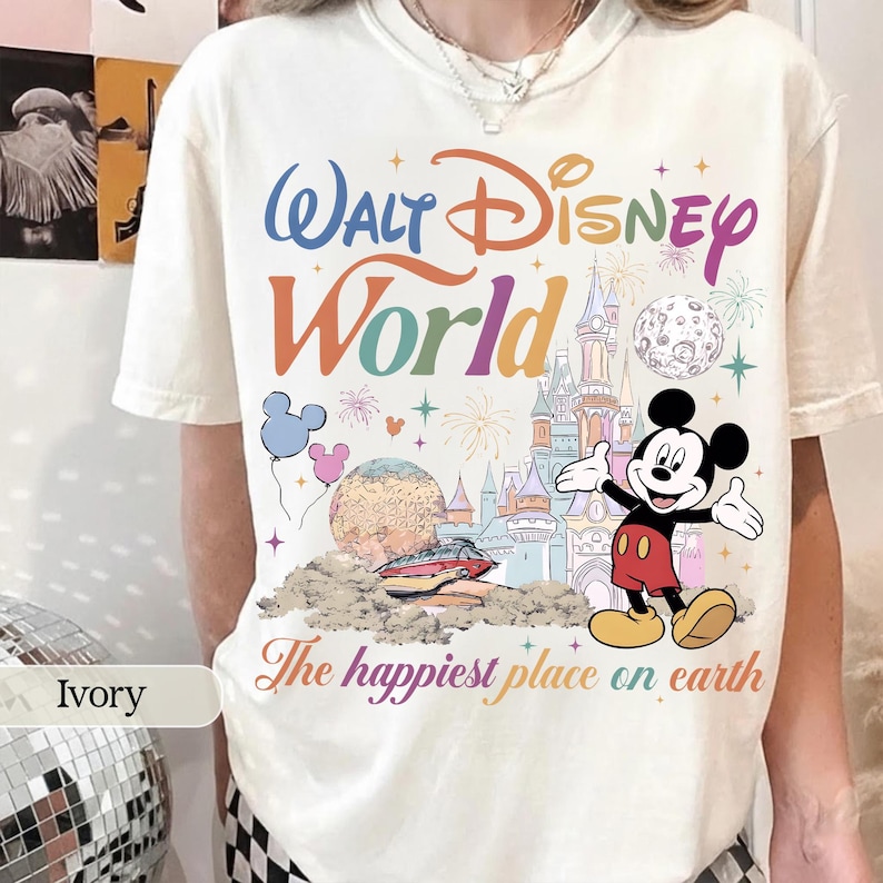 May include: Ivory-colored t-shirt featuring a colorful "Walt Disney World" graphic with Mickey Mouse, a castle, and the phrase "The happiest place on earth." The design includes balloons, fireworks, and a planet.