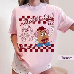 Puede incluir: Camiseta rosa claro con un dise&ntilde;o de Mr. Potato Head y el texto "Mr. Potato Head 1995". El dise&ntilde;o incluye un patr&oacute;n de cuadros rojos oscuros. La camiseta tiene mangas cortas.