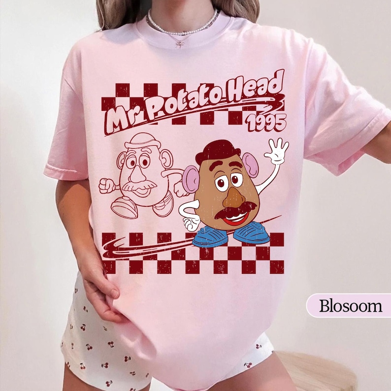 Puede incluir: Camiseta rosa claro con un dise&ntilde;o de Mr. Potato Head y el texto "Mr. Potato Head 1995". El dise&ntilde;o incluye un patr&oacute;n de cuadros rojos oscuros. La camiseta tiene mangas cortas.