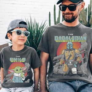K&ouml;nnte beinhalten: Zwei anthrazitfarbene T-Shirts mit Grafikdrucken. Das kleinere Shirt zeigt eine gr&uuml;ne Kreatur mit dem Text THE CHILD. Das gr&ouml;&szlig;ere Shirt zeigt einen gepanzerten Charakter mit dem Text THE DADALORIAN. Beide haben eine rote Sonne als Hintergrund.