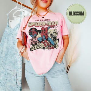Op de afbeelding: Een lichtroze t-shirt met een vintage Spider-Man-afbeelding. Het ontwerp bevat de woorden "THE AMAZING SPIDER-MAN", "SINTO ACTION" en "NEW YORK 1962". De afbeelding toont Spider-Man in actie.