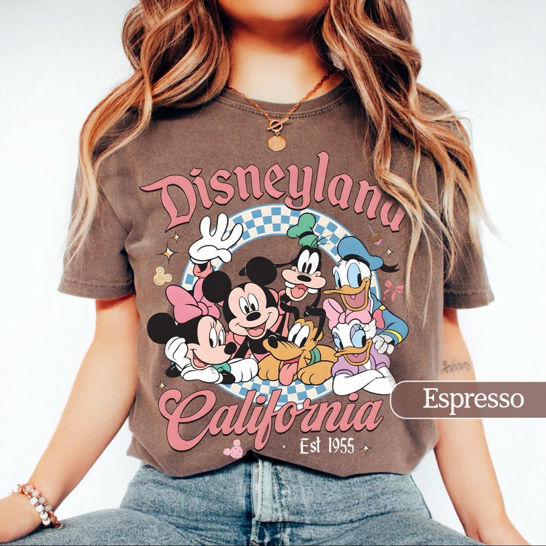 May include: Espresso-colored t-shirt featuring a graphic of Disney characters, including Mickey and Minnie Mouse, Goofy, Donald and Daisy Duck. The text "Disneyland California" is displayed in pink script, with "Est 1955" below.
