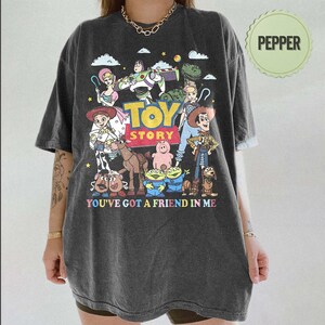 May include: Dark gray t-shirt featuring a colorful graphic of Toy Story characters, including Woody, Buzz Lightyear, and others. The text "TOY STORY" and "YOU'VE GOT A FRIEND IN ME" are also printed on the shirt. The shirt has a relaxed fit.