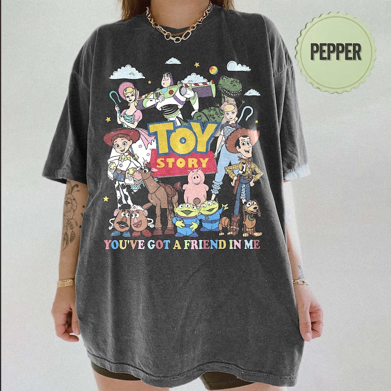 May include: Dark gray t-shirt featuring a colorful graphic of Toy Story characters, including Woody, Buzz Lightyear, and others. The text "TOY STORY" and "YOU'VE GOT A FRIEND IN ME" are also printed on the shirt. The shirt has a relaxed fit.