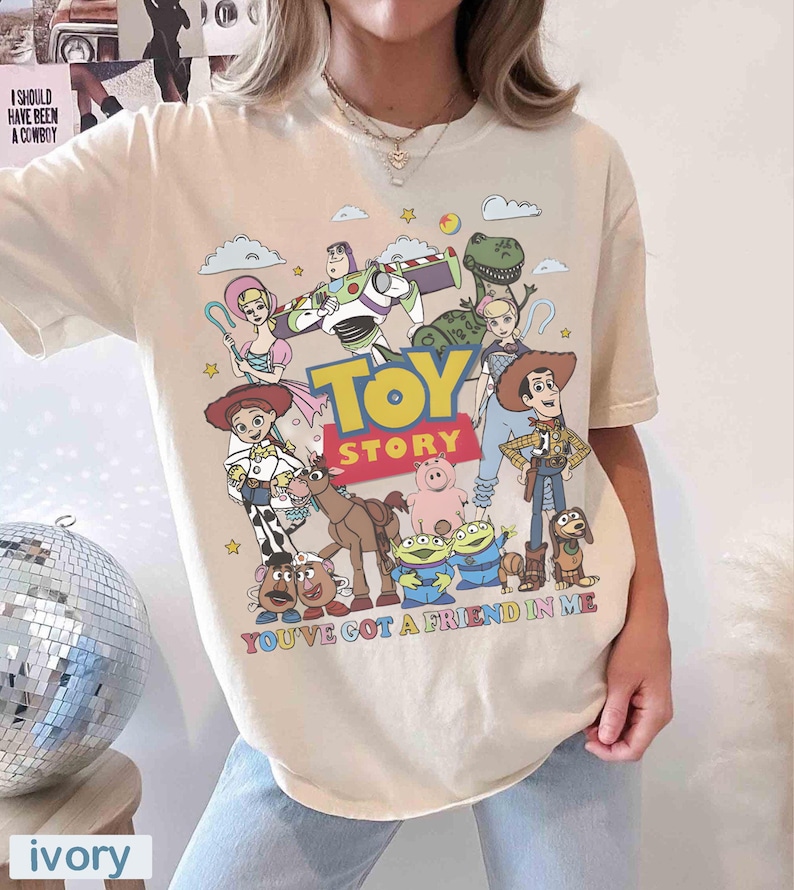 May include: Ivory-colored t-shirt featuring a colorful graphic of Toy Story characters, including Woody, Buzz Lightyear, and others. The text "TOY STORY" is prominently displayed, with "YOU'VE GOT A FRIEND IN ME" below.