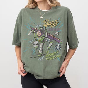 Puede incluir: Camiseta verde oliva con un dise&ntilde;o de Buzz Lightyear y las frases "Buzz Lightyear" y "To Infinity and Beyond". El dise&ntilde;o incorpora un tema espacial con un planeta y estrellas.