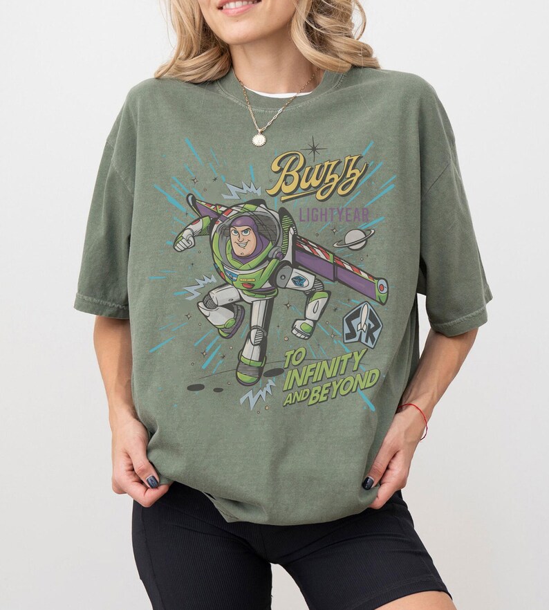 Puede incluir: Camiseta verde oliva con un dise&ntilde;o de Buzz Lightyear y las frases "Buzz Lightyear" y "To Infinity and Beyond". El dise&ntilde;o incorpora un tema espacial con un planeta y estrellas.
