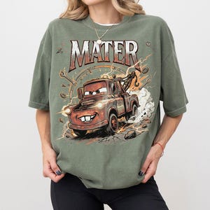 May include: Olive green t-shirt featuring a cartoon image of Mater from the movie Cars. The graphic includes the word "MATER" in a distressed font, along with tools and lightning bolts. The image depicts Mater with a big smile, driving fast.