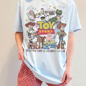 May include: Light blue t-shirt featuring a colorful graphic of Toy Story characters. The design includes Woody, Buzz Lightyear, and other characters from the movie, with the text "TOY STORY" and "YOU'VE GOT A FRIEND IN ME".