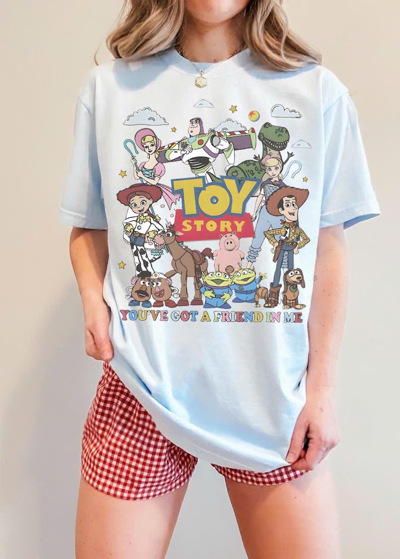 May include: Light blue t-shirt featuring a colorful graphic of Toy Story characters. The design includes Woody, Buzz Lightyear, and other characters from the movie, with the text "TOY STORY" and "YOU'VE GOT A FRIEND IN ME".