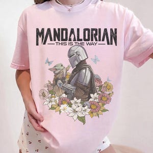 May include: Pink t-shirt featuring a graphic of The Mandalorian and Grogu surrounded by flowers and butterflies. The text "MANDALORIAN - THIS IS THE WAY" is printed across the top in black.