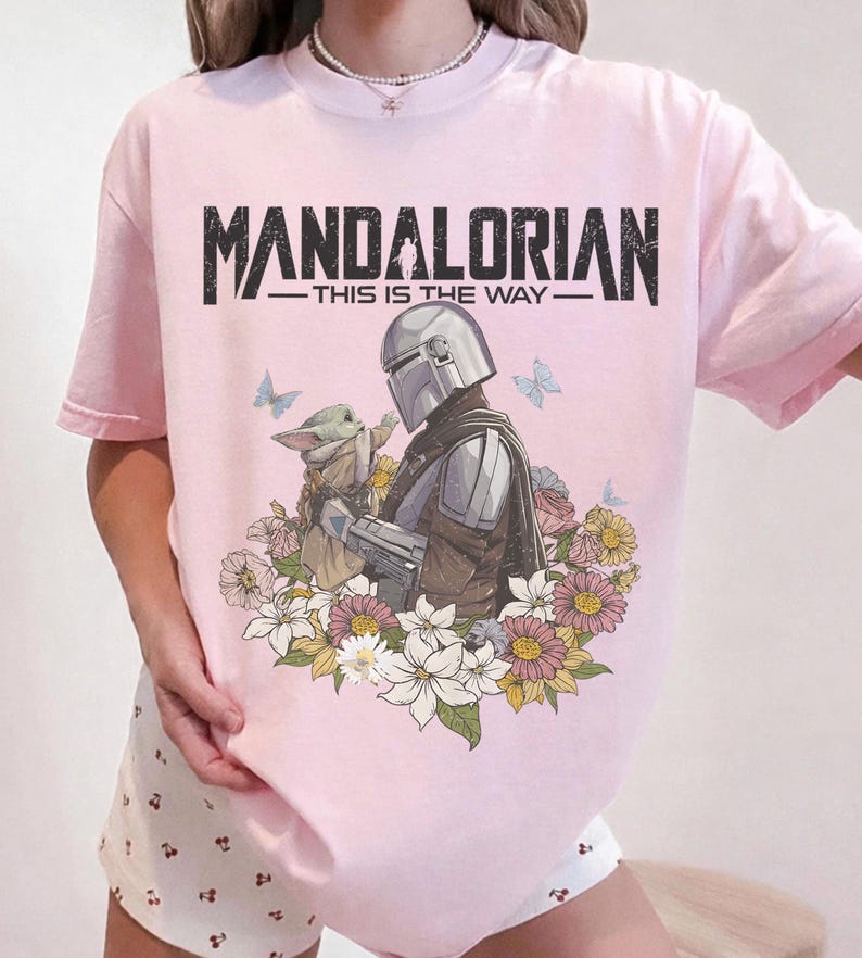 May include: Pink t-shirt featuring a graphic of The Mandalorian and Grogu surrounded by flowers and butterflies. The text "MANDALORIAN - THIS IS THE WAY" is printed across the top in black.
