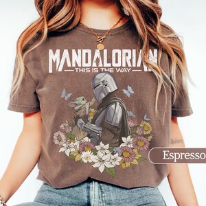 May include: Espresso-colored t-shirt featuring a graphic of The Mandalorian and Grogu surrounded by flowers and butterflies. The text "MANDALORIAN - THIS IS THE WAY" is printed across the top in white.