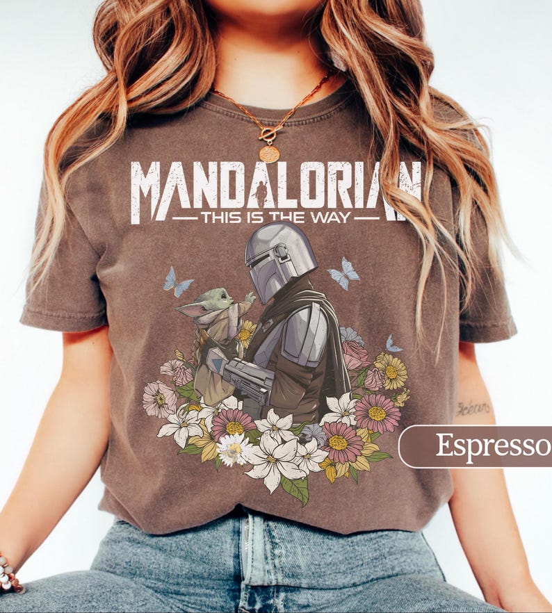 May include: Espresso-colored t-shirt featuring a graphic of The Mandalorian and Grogu surrounded by flowers and butterflies. The text "MANDALORIAN - THIS IS THE WAY" is printed across the top in white.