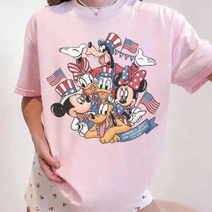 May include: A light pink t-shirt featuring a graphic of Disney characters Mickey Mouse, Minnie Mouse, Donald Duck, Daisy Duck, Goofy, and Pluto, all in patriotic attire, holding American flags. The text "AMERICA 250th USA ANNIVERSARY 1776-2026" is on a blue banner.