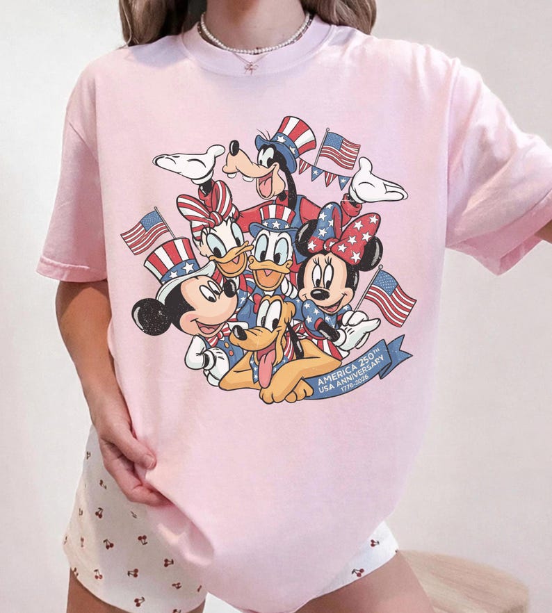 May include: A light pink t-shirt featuring a graphic of Disney characters Mickey Mouse, Minnie Mouse, Donald Duck, Daisy Duck, Goofy, and Pluto, all in patriotic attire, holding American flags. The text "AMERICA 250th USA ANNIVERSARY 1776-2026" is on a blue banner.