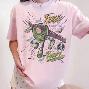 Puede incluir: Camiseta rosa claro con un dise&ntilde;o de Buzz Lightyear en su traje espacial, con las palabras "Buzz Lightyear" y "To Infinity and Beyond". El dise&ntilde;o incluye un planeta y explosiones de estrellas.