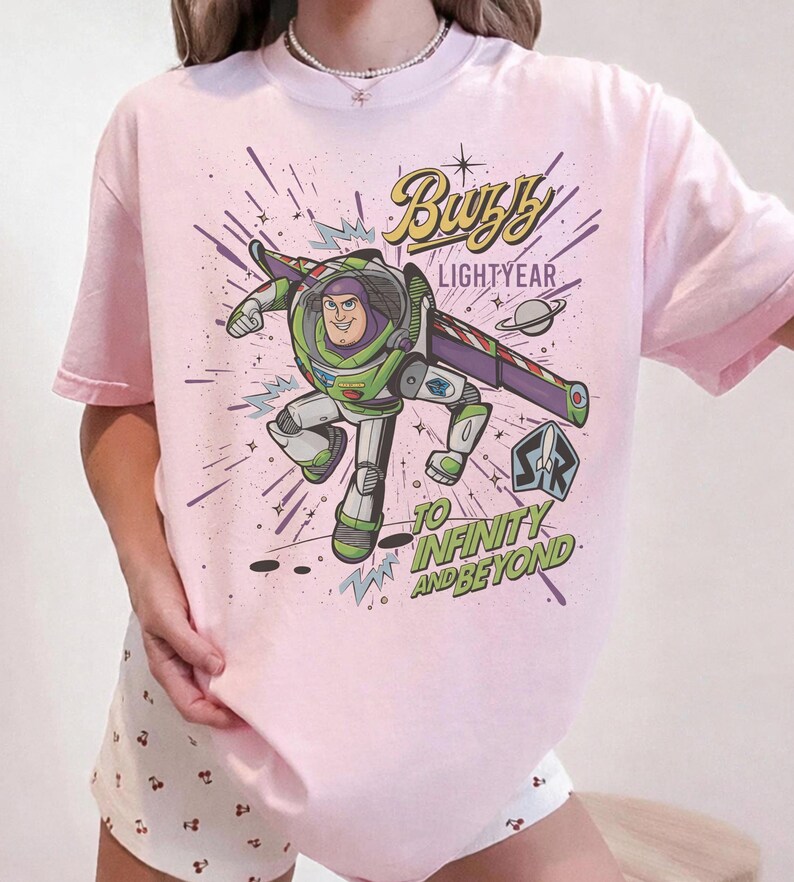 Puede incluir: Camiseta rosa claro con un dise&ntilde;o de Buzz Lightyear en su traje espacial, con las palabras "Buzz Lightyear" y "To Infinity and Beyond". El dise&ntilde;o incluye un planeta y explosiones de estrellas.